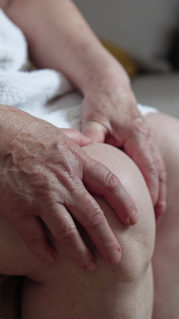 Close-up of experienced hands performing therapeutic massage on client's knee. Soft lighting creates intimate wellness atmosphere. Perfect for healthcare, spa, and senior care marketing.の素材