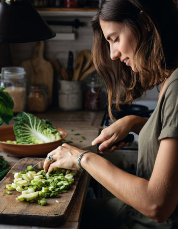 Concentrated brunette preparing healthy meal, dicing celery on wooden board. Warm natural lighting creates intimate cooking atmosphere. Perfect for wellness, nutrition, lifestyle content.の素材