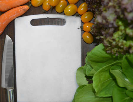 empty white cutting board in the middle of lettuce, mustard green, knife, orange carrots and yellow...