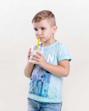Charming little boy in a blue t-shirt drinks milk from a yellow straw. A cute little boy is holding a glass of milk. Adorable little boy is drinking fresh milk on gray background.の写真素材