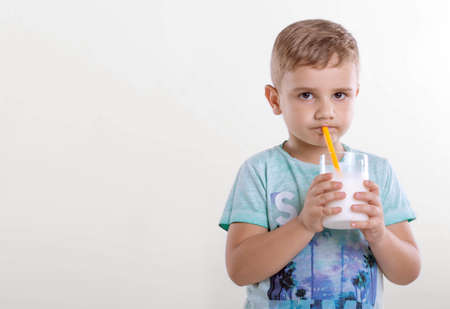 Portrait of a nice little boy in a blue T-shirt,  with a glass of milk and yellow straw. A cute little kid drinking a milk from a yellow straw on gray background.の写真素材