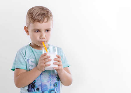 Pretty little boy in the blue t-shirt is drinking milk from a yellow straw on a gray background. Beautiful child with glass of milk on gray background. Healthy eating little boy is drinking milk from a yellow straw.の写真素材