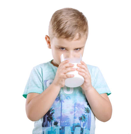 Adorable little kid is holding a glass of organic milk and drinking fresh milk isolated on awhite background. Close-up portrait of a cute little boy in a blue t-shirt with a full glass of milk.の写真素材