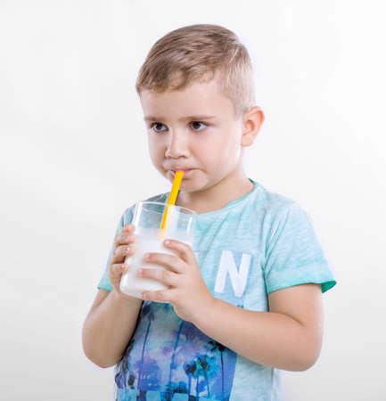 Adorable little boy holding a glass of organic milk and drinking fresh milk on gray background. Close-up portrait of a cute little kid in a blue T-shirt with a full glass of milk from yellow straw.の写真素材