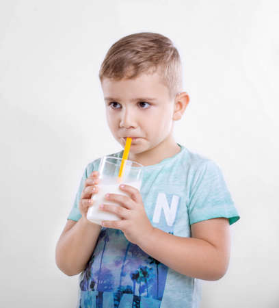 Pretty little boy is drinking milk in a glass from a yellow straw. Close-up portrait of cute little kid is drinking milk from yellow straw in the morning on gray background. Concept of healthy living, food  drink, family.の写真素材