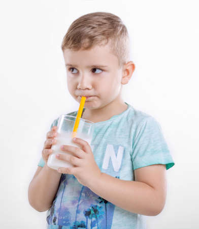 Cute little boy holding a glass of milk and drinking fresh milk. A little kid standing in blue shirt and drinking milk in a glass from a yellow straw. A nice little kid with milk. Healthcare.の写真素材