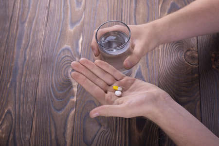A man holds two pills and a glass of water in the hands of a wooden brown table. The guy drinks pills. Healthy generation. Healthy youth.の写真素材