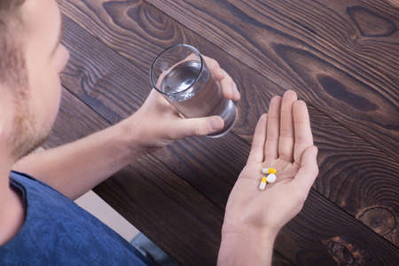 A young guy with a beard in a blue T-shirt is holding three tablets of different colors and a glass of water against the background of a wooden brown table. Healthy generation. Healthy youth.の写真素材