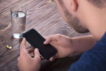 A young guy in a blue T-shirt sits at a wooden brown table and holds a phone in his hands.  There is a glass of water, one white pill lies, and the other is a yellow tablet on the wooden table.の写真素材
