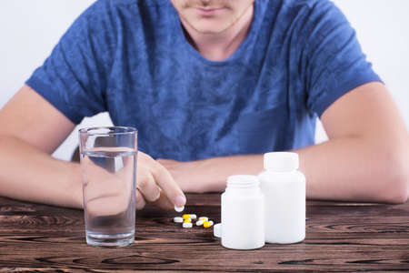 A young guy in the blue T-shirt sits at a wooden brown table and holds a pill in his hand. There is a glass of water on the table, two boxes of medicines and white and yellow tablets on the table.の写真素材
