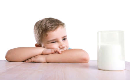 A little adorable thoughtful boy in a blue T-shirt lies on the bright wooden table and wonders about the taste of nutritious milk standing right next to him in a big glass.の写真素材