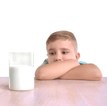 An adorable little boy in a blue t-shirt is lying on a big bright wooden desk and looking at a huge glass full of healthy and fresh milk, isolated on a white background.の写真素材