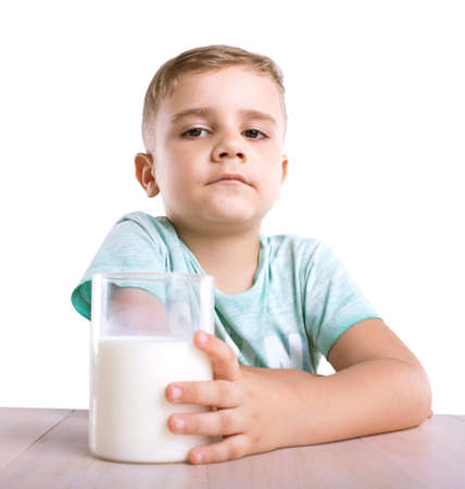 A curious and nice little boy in a blue t-shirt holds a glass of milk and looks cute, isolated on a white background.  Close-up portrait of a handsome little kid with a glass full of nutritious milk.の写真素材