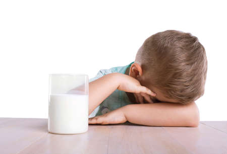 A tired adorable small kid with blonde hair lies on the light brown classic table with a glass of nutritious and tasty milk, isolated on a white background.の写真素材