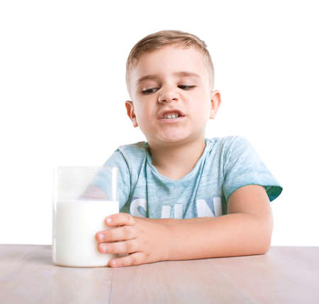 A lovely and adorable little kid is sitting next to the glass of fresh milk. A cute little boy does not want to drink milk, isolated on a white background. Small boy isn't pleased with his glass of a healthful milk.の写真素材