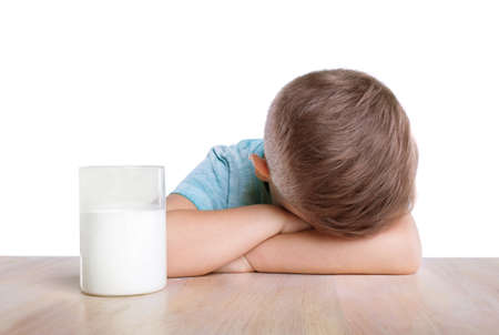 A big spacious glass full of organic and tasty white milk and a sad charming blonde boy in a blue T-shirt. The Charming little boy is lying on his crossed hands on the classic table.の写真素材