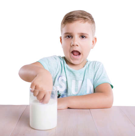 Charming little blond boy in a blue t-shirt is touching a nutritious and tasty milk in a big transparent glass on a light brown classic table, isolated on a white background.の写真素材