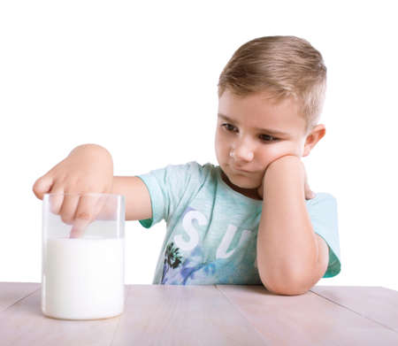 A thoughtful small blonde boy in a bright blue T-shirt is sitting right next to the glass of milk. The cute baby boy is dipping his tiny finger in the organic milk drink, isolated on white background.の写真素材