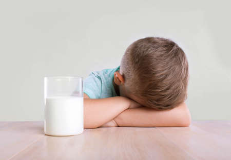 A tiny cute little boy in a blue t-shirt is sitting near to the transparent glass full of organic milk. The handsome little boy with a glass full of tasty milk, on a gray background.の写真素材