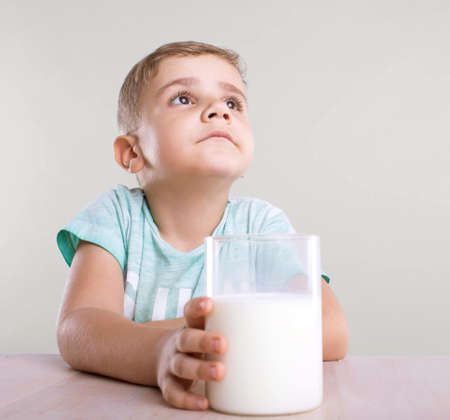 Thoughtful and little boy in a blue t-shirt is dreaming and holding a huge glass of nutritious and organic milk on a polished light brown table, on a gray background.の写真素材