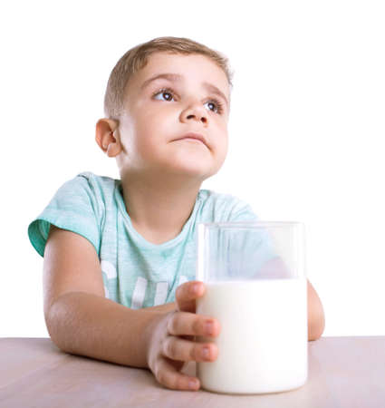 A nutritious milk in a massive glass on a light brown wooden table. A little cute boy in a blue t-shirt holds the glass, dreaming about a tasty of milk and looks up, isolated on a white background.の写真素材