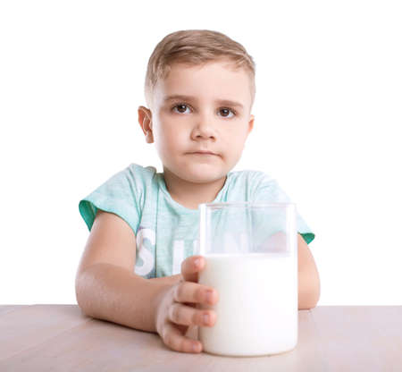 An adorable and careful little boy in a blue T-shirt, with a transparent glass of delicious white milk in a hand, sits next to the polished brown table, isolated on a white background.の写真素材