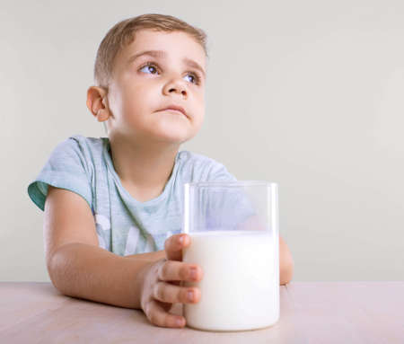 An enormous glass full of healthful tasty milk in a hand of an adorable and sad tiny kid with a blond hair, in a blue T-shirt, isolated on a gray background.の写真素材