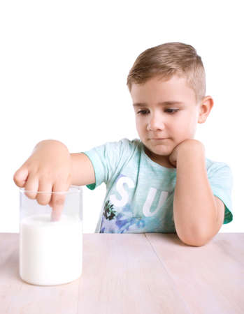 Close-up portrait of a little boy with blonde hair and in a pretty blue t-shirt with a glass full of fresh milk. A handsome little boy is dipping his tiny finger in milk isolated on a white backgroundの写真素材
