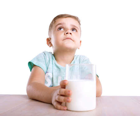 A wonderful little kid with a golden hair sits close to the light brown classic table and holds a glass full of tasteful and white milk, looks very cute, isolated on a white background.の写真素材
