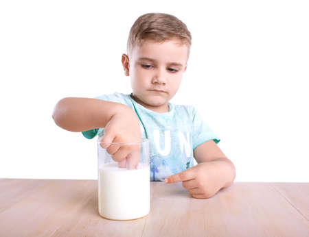 A cute and curious little boy in a blue t-shirt looks at milk and dips his small hand in it and points at the glass beaker with milk on a light brown wooden table, isolated on a white background.の写真素材