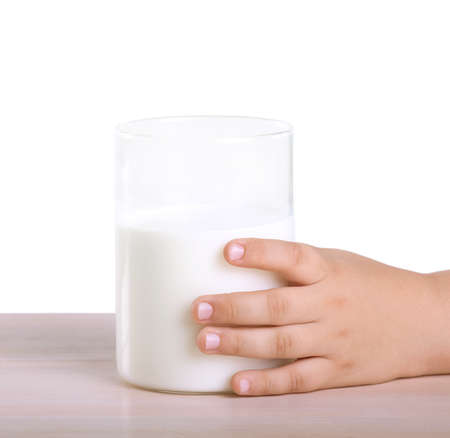 A small hand of a little child is holding a large glass of organic milk, which stands on a wooden table isolated on a white background.の写真素材