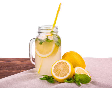 A glass jar with a yellow straw and a cooling mojito with bright green mint leaves and a yellow lemon on a brown table, isolated on a white background. Tasty fresh cocktail with ripe and bright yellow lemon and green mint.の写真素材