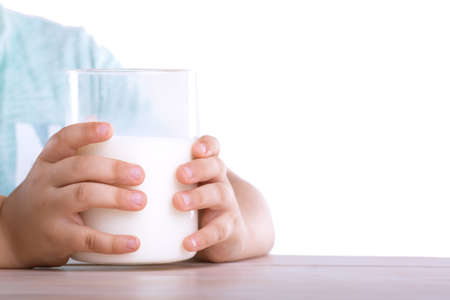 The small fingers of a little child in a blue T-shirt are holding a large glass of organic milk, which stands on a wooden table isolated on a white background.の写真素材