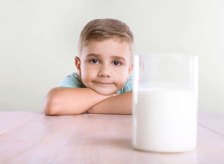 At wooden table sits a cute and attractive little boy in a blue T-shirt with a glass of organic milk isolated on a white background. Close-up portrait of a happy little boy with a glass of fresh milkの写真素材