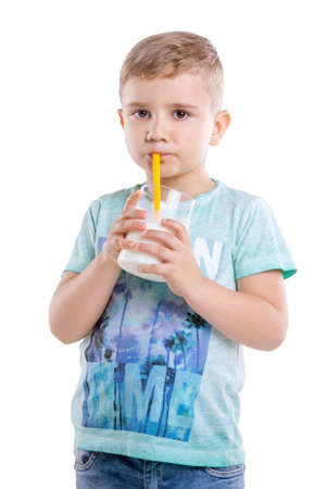 A beautiful little boy in a blue T-shirt is drinking fresh and organic milk from a yellow straw. Adorable little child with a glass of milk  and yellow straw, isolated on a white background.の写真素材