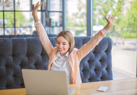 Fantastic lady sits on the sofa, looks on the laptop and happy. Portrait of amazing and joyfull business woman in white blouse. A nice business woman is smiling. The laptop and phone are on a table.の写真素材