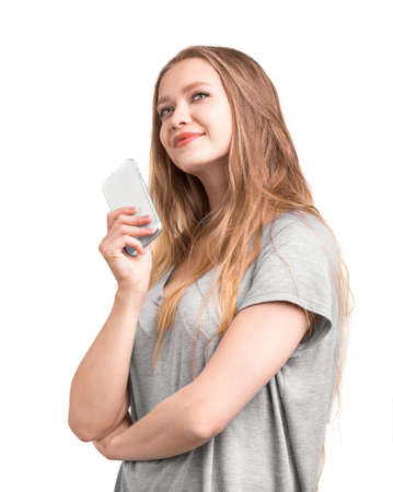 A beautiful girl in a gray T-shirt is holding a phone and dreaming. Close-up portrait of a thoughtful, charming and fashionable girl stands and thinks who to call on the phone.の写真素材