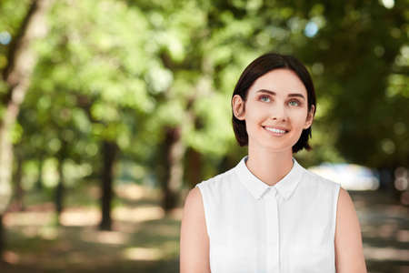 A cheerful young woman on a natural background. A beautiful brunette business lady smiling and walking in a forest in summer. A confident, happy and elegant girl outdoor. A beautiful girl looking up.の写真素材