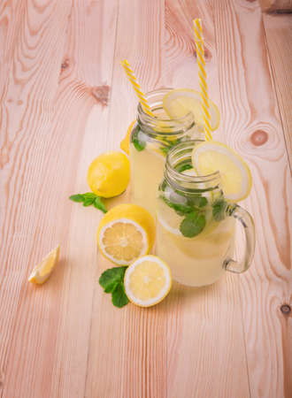 Summer smoothie from ripe yellow lemons, fresh mint and mineral water in two huge masone jars with a yellow straws and with pieces of lemons at the top of the glasses on a light wooden table.の写真素材