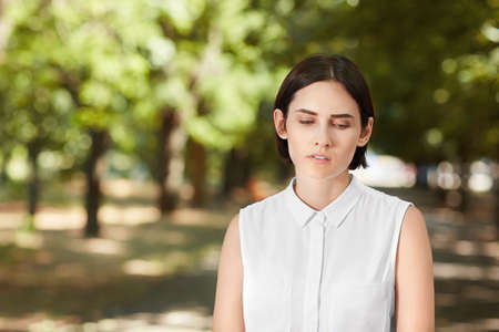 A beautiful successful girl standing in a park. A thoughtful young woman is looking down in an elegant blouse. Pretty brunette girl is walking outdoors. Business lady in a park.の写真素材