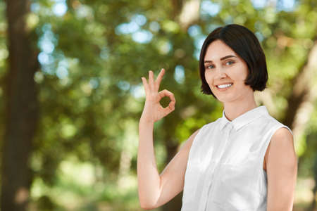 A cheerful young lady on a bright light natural background. A brunette attractive girl making an "OK" sign. Health, confidence, beauty, freshness concept. Pretty girl having fun in a forest.の写真素材