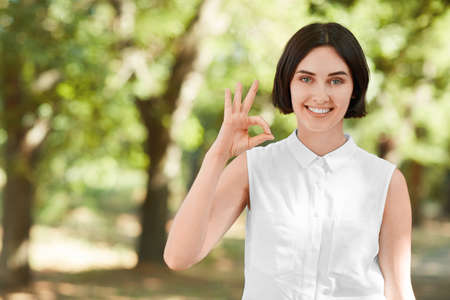 A smiling confident woman in a light white shirt on a bright green, natural background. Happy girl making an "OK" sign. Portrait of a healthy young joyful lady in a park.の写真素材