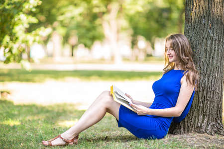 A cute, perfect, cheerful, and wonderful girl in a bright blue dress sits on a green grass near a tree. A beautiful lady is reading an interesting book and smiling. Beautiful girl with book.の写真素材