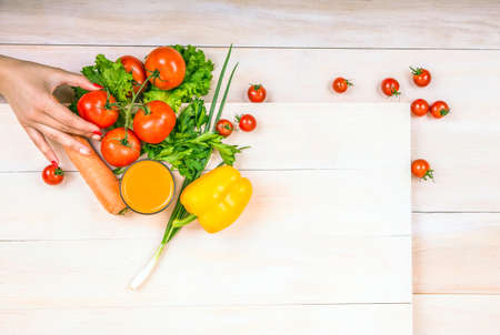 Close-up food ingredients on a light background. Tasteful vegetables on a spacious wooden table. A hand holding a fresh red tomato, laying next to the lettuce. A glass full of orange juice.の写真素材