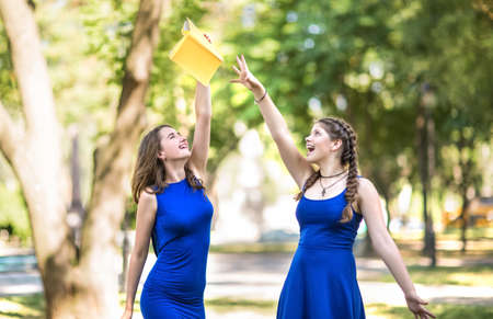 Two charming gorgeous, beautiful girls in blue dresses in the park. A beautifully looking girl with a left holds a  book in her hands. An elegant lady with the right has beautiful pigtails. Cute girlsの写真素材