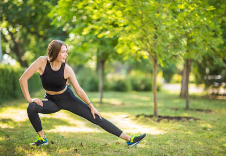 A woman with beautiful long blond hair is doing stretching in the green park in the morning. The charming girl is dressed in a black top, black leggings and black trainers with green accents.の写真素材