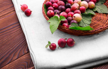 A brown wooden basket with multi-colored gooseberries on white fabric on a dark brown background. Colorful red berries full of nutritious vitamins. Close-up ripe fresh red berries. Delicious berries.の写真素材