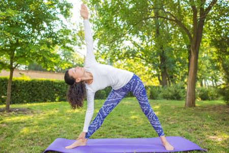 Sports girl doing yoga in a summer park.  Relaxing exercises. Concentrated flexible girl on a green natural background. Skinny girl practicing active training. The lady on a purple mat.の写真素材