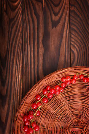 A close-up picture of a ripe and nutritious red currant in an upright position. A colorful partial composition is in the lower right corner of a brown basket with juicy red currant on a wooden table.の写真素材