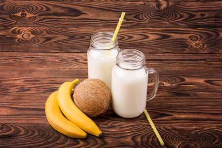 Delicious fruits and jars full of milk on a bright wooden background. Mason jars with yellow straws full of nutritious milk. Fresh tropical fruits. Beautiful bananas and a hawaiian coconut.の写真素材